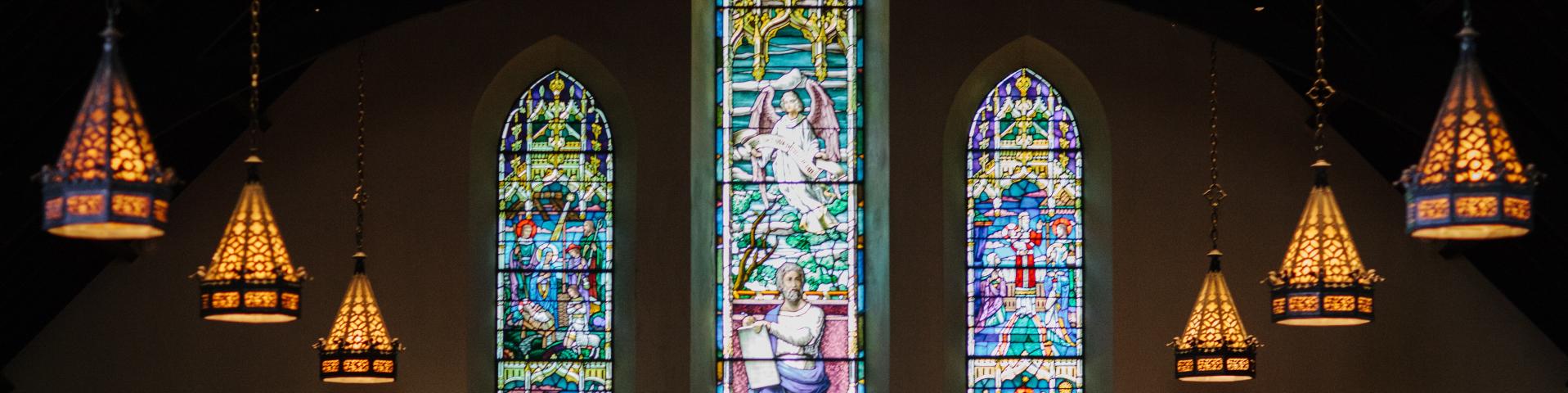 Dark church with three stained glass windows and 3 golden stained glass chandeliers hanging on each side