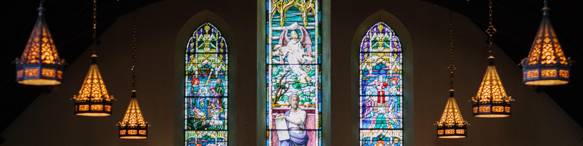 Dark church with three stained glass windows and 3 golden stained glass chandeliers hanging on each side