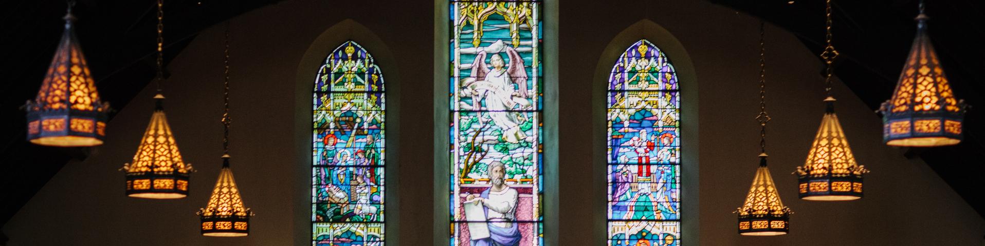 Dark church with three stained glass windows and 3 golden stained glass chandeliers hanging on each side