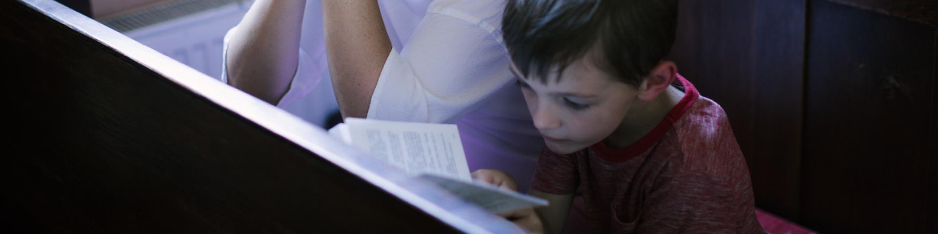 Little boy sitting in a pew praying with a book in his hands. Mother with rosary looking over his shoulder at book
