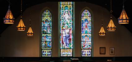 Dark church with three stained glass windows and 3 golden stained glass chandeliers hanging on each side